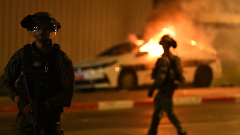 Agentes de policía patrullan las calles de Lod, mientras un patrullero arde al fondo. 